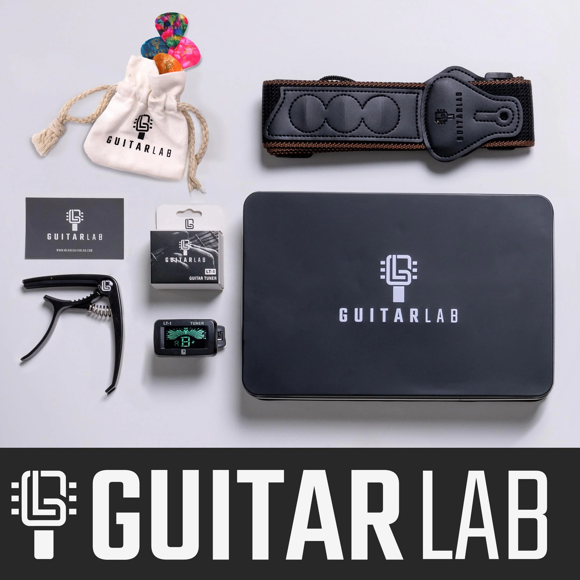 Pudełko prezentowe z akcesoriami gitarowymi [marki Guitar Lab], metalowa puszka zawiera pasek do gitary, kapodaster, elektroniczny tuner i puszkę, idealne dla miłośników gitary i uczących się gry